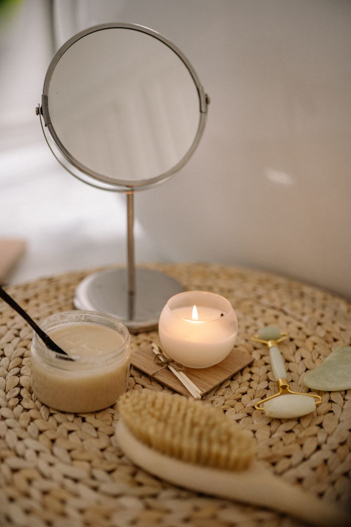A calming spa scene with a lit candle, mirror, brush, and jade roller on a woven mat.